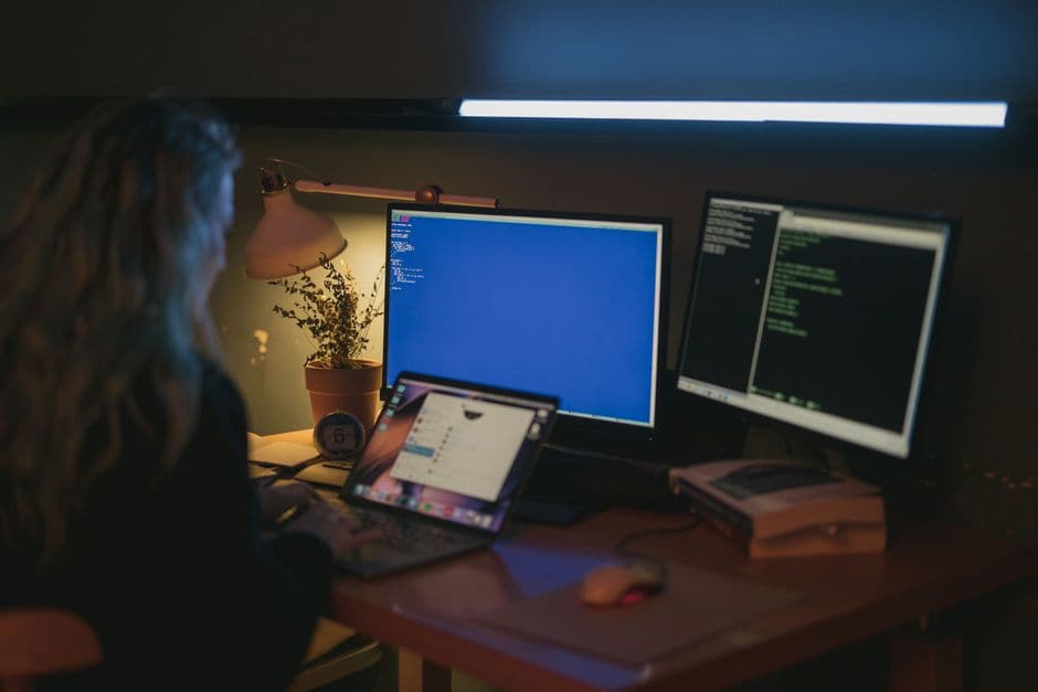 暗い部屋で人物がノートパソコンと2台の外部モニターを使ってコードを確認している写真
