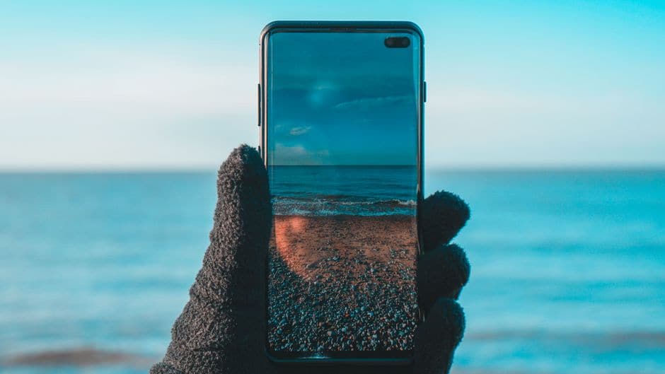 海辺で持ち上げたスマートフォンの画面に、目の前の海と砂浜の景色が映り込んでいる写真