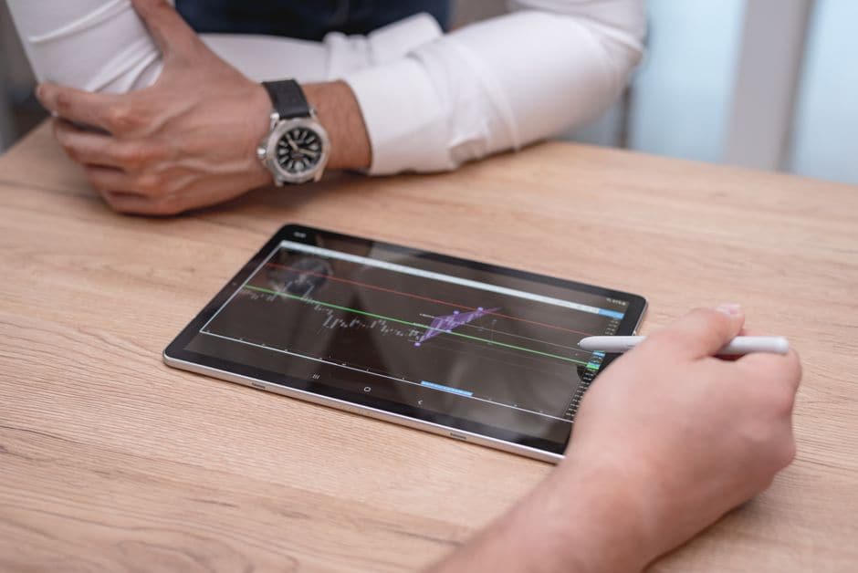 木製のデスクの上でスタイラスペンを使い、金属色のタブレットでチャートを操作する手元の接写