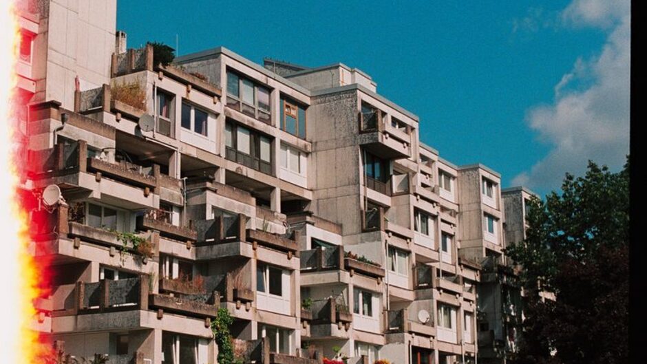 青空の下に立つ幾何学的な集合住宅。画面左端に赤い光が差している
