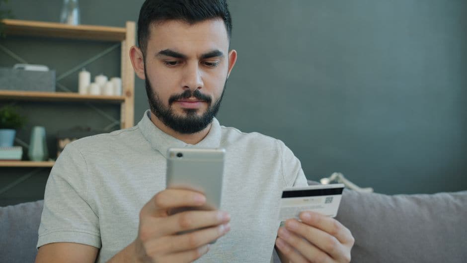 男性がクレジットカードのような薄いカードを片手に持ち、もう片方の手のスマートフォンを見ながら操作している室内の写真