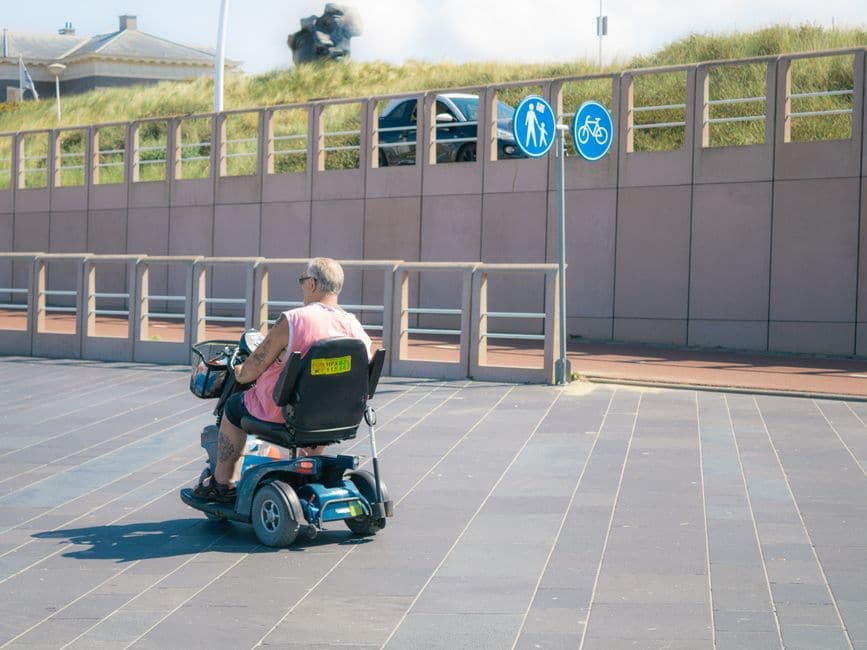 高齢の男性が青い電動モビリティスクーターに座り、歩行者・自転車の標識がある遊歩道を走っている写真